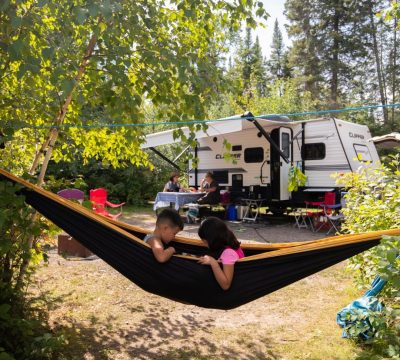 Two children in a hammock with an camping trailer and their parents in the background, showcasing the quality time Whiteshell Campgrounds provide.
